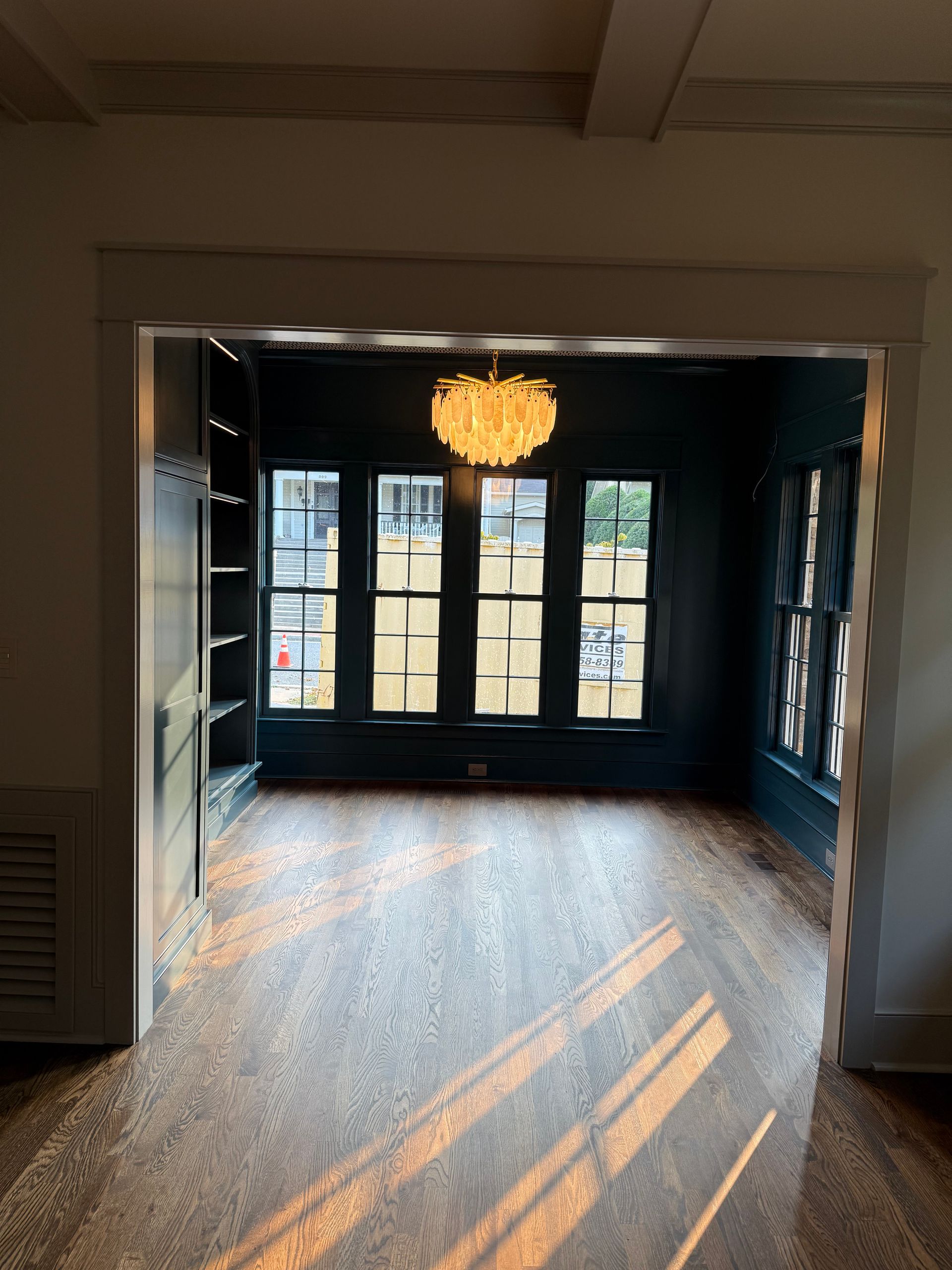 Room with dark blue walls, built-in shelving, large windows, and a decorative chandelier, viewed from an adjacent room.