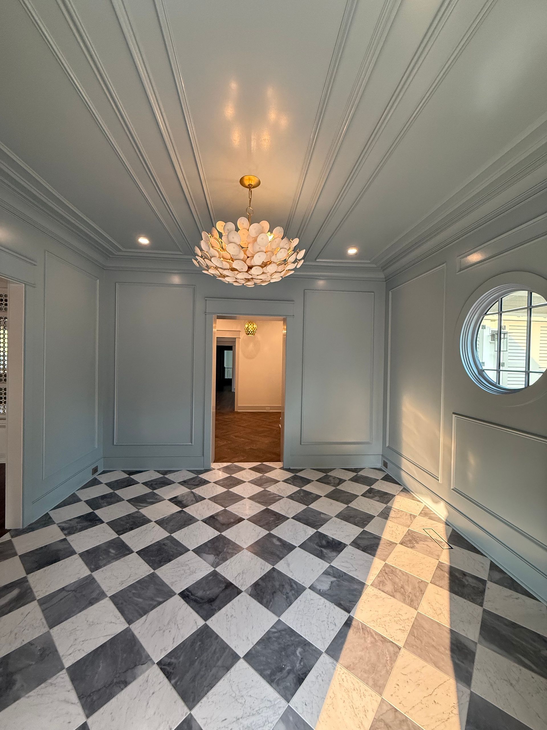 Room with a checkered floor, light blue walls, and a floral chandelier. A circular window is visible.