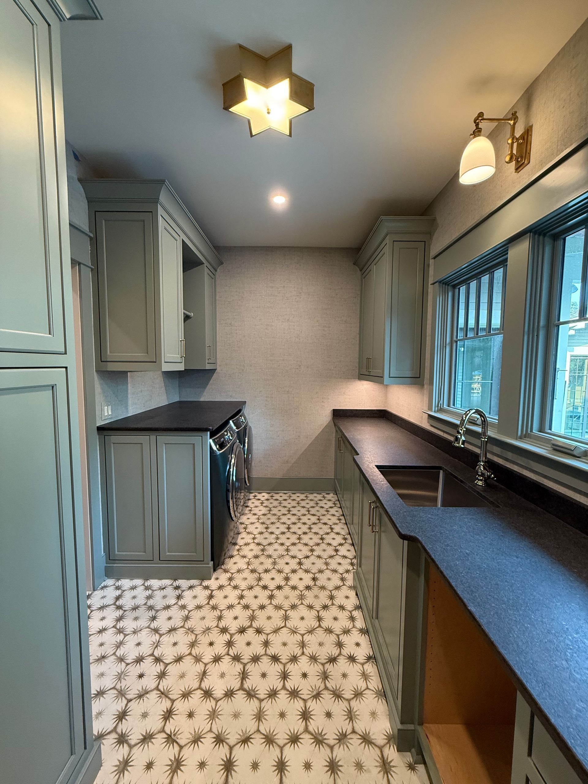 Narrow kitchen with gray cabinets, black countertops, patterned tile floor, and a window.