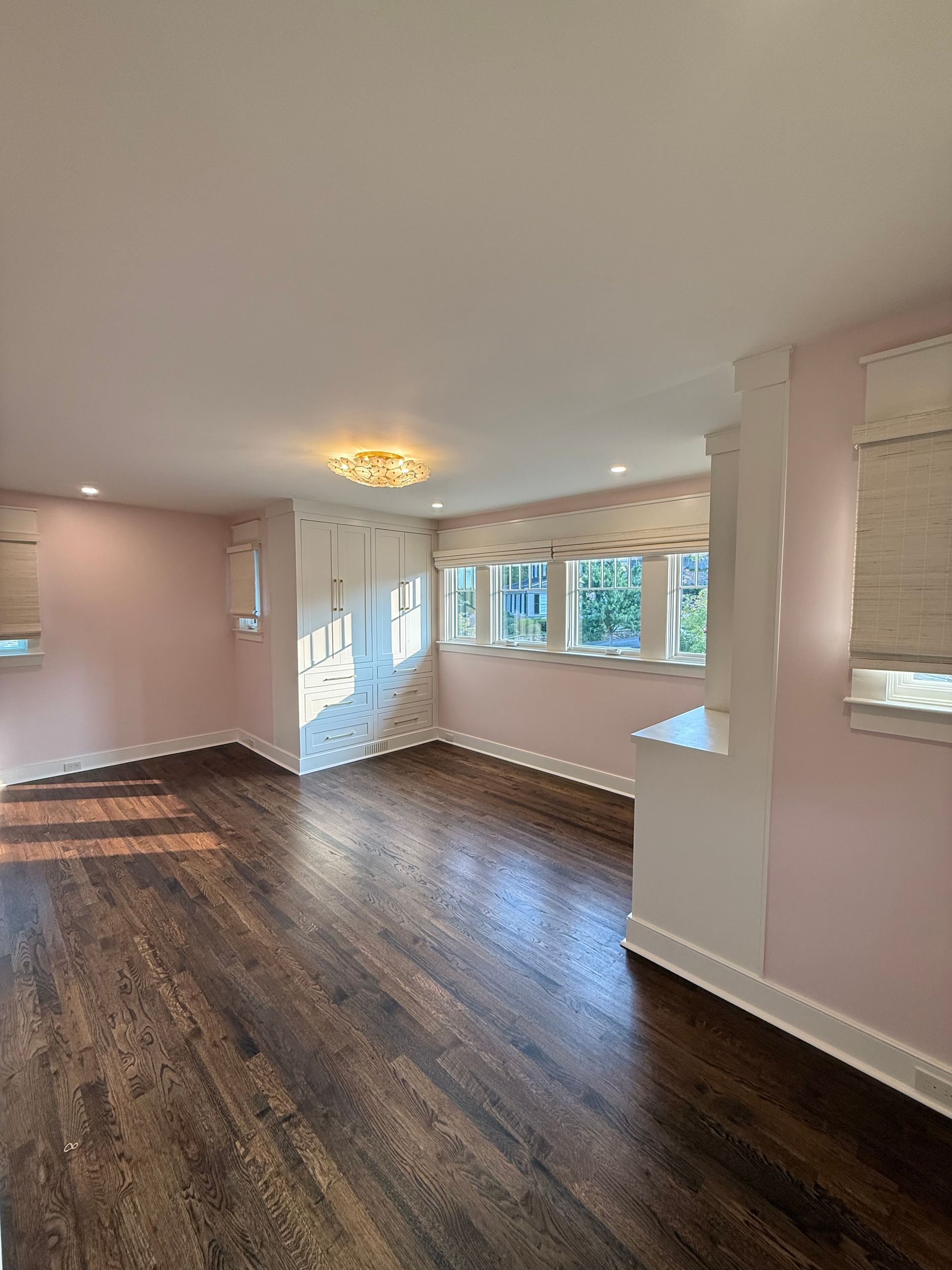 Empty room with dark wood floors, pink walls, and white trim. Windows with light shades.