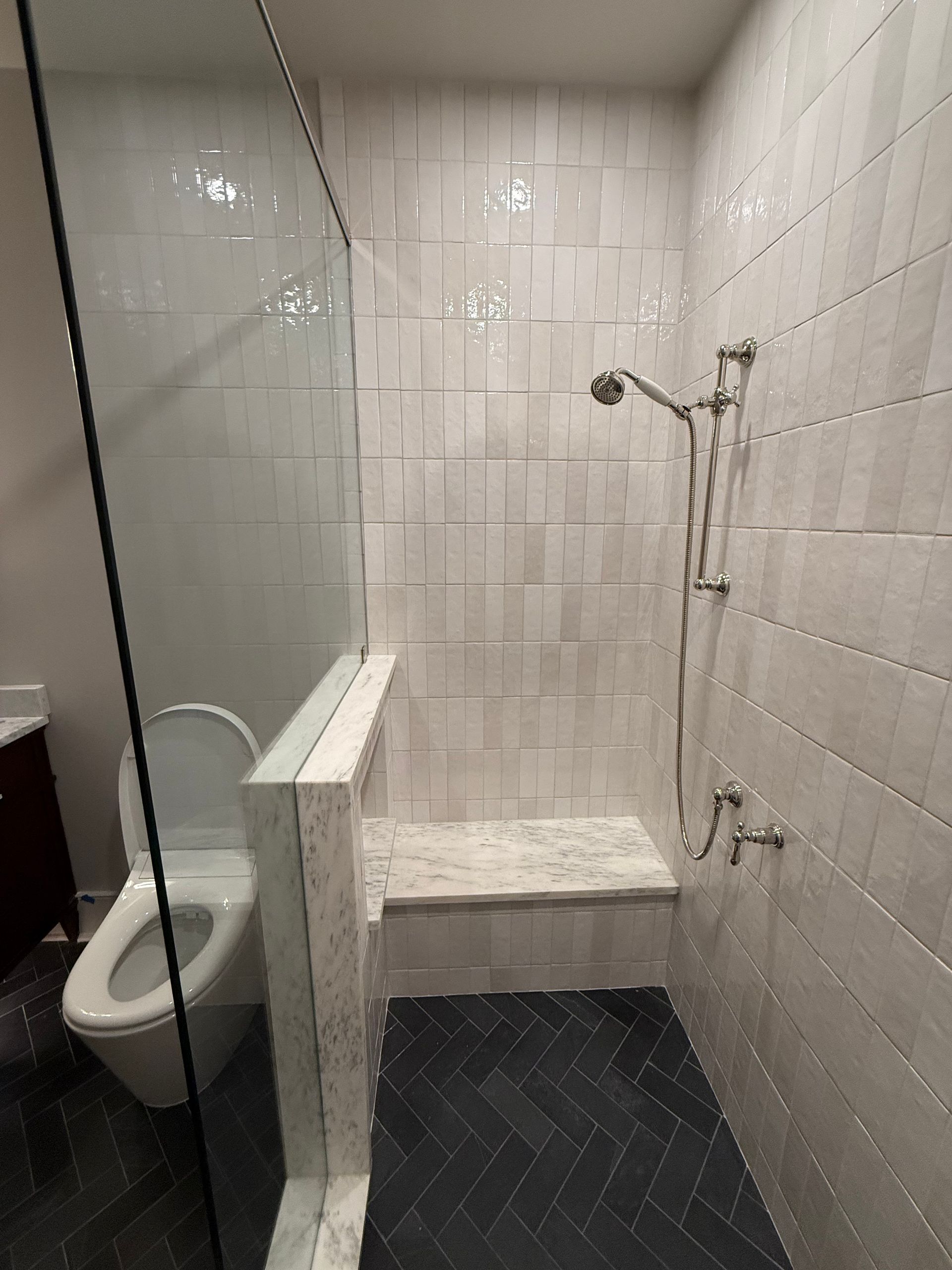 Bathroom with white tile walls, glass shower door, and a built-in shower seat. The floor is dark herringbone tile.