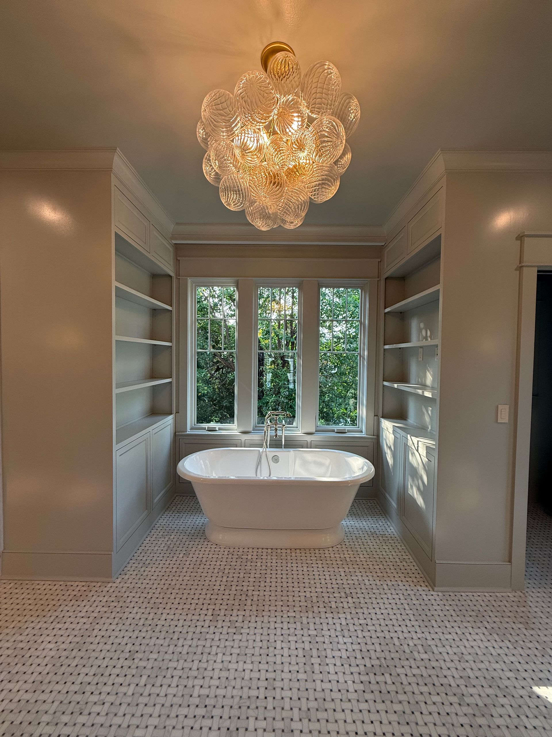 Elegant bathroom with a free-standing tub, window, shelves, and a bubble chandelier. White and neutral tones.