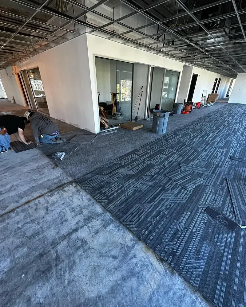 Workers installing gray carpet tiles in an unfinished office space. The room has white walls and sliding glass doors.