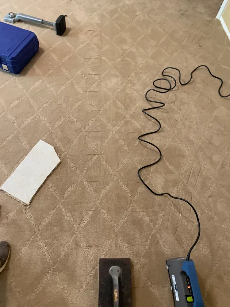 Carpet being installed, tools visible. Tan diamond-patterned carpet, blue tool case, black power cord.