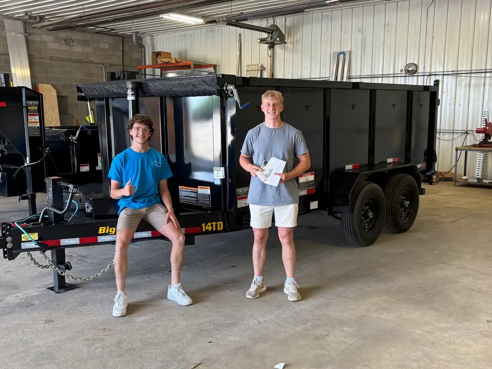 Two young men are standing next to a dump trailer in a garage.