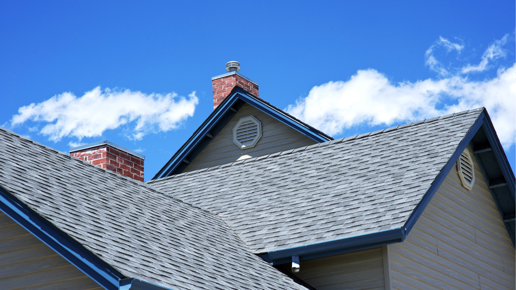 Grey shingled house roof with a brick chimney and blue trim against a bright blue sky with clouds.