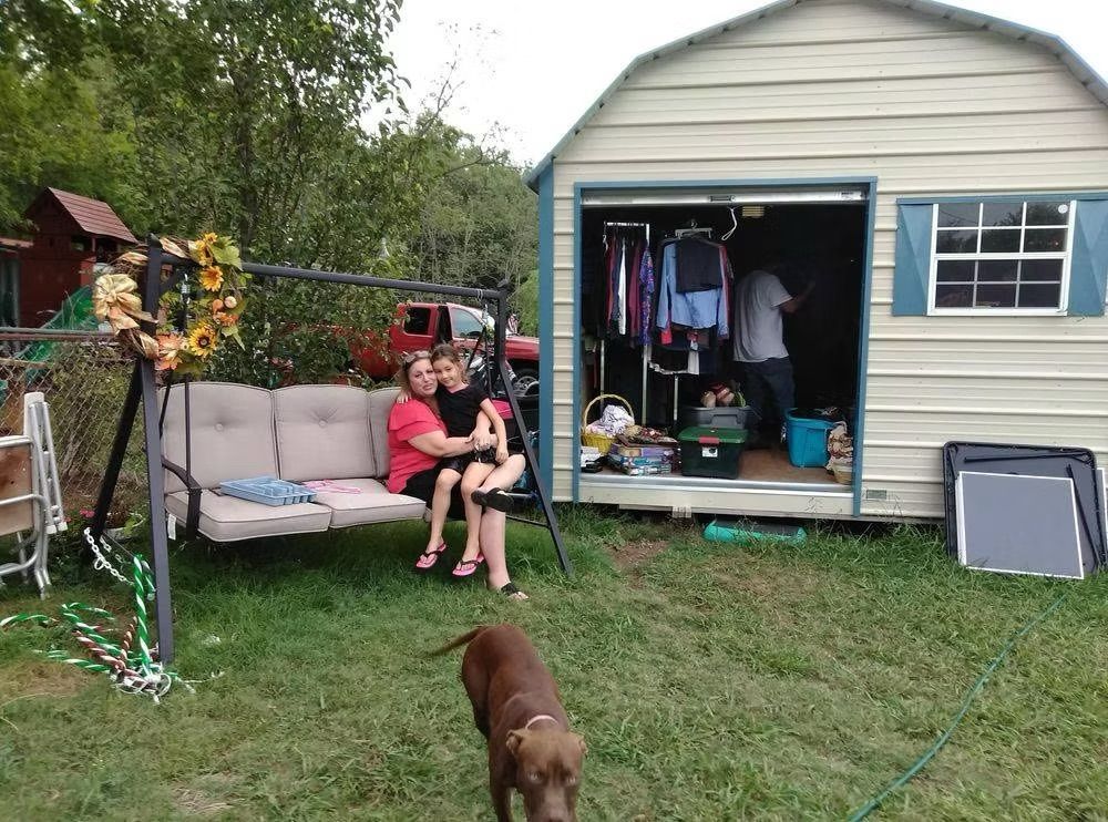 Two people sit on a swing in a yard, a dog in the foreground, shed with open door behind them.
