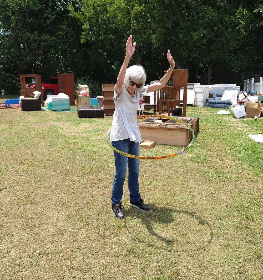 Woman in jeans and a t-shirt hula-hooping outside with arms raised, furniture in the background.