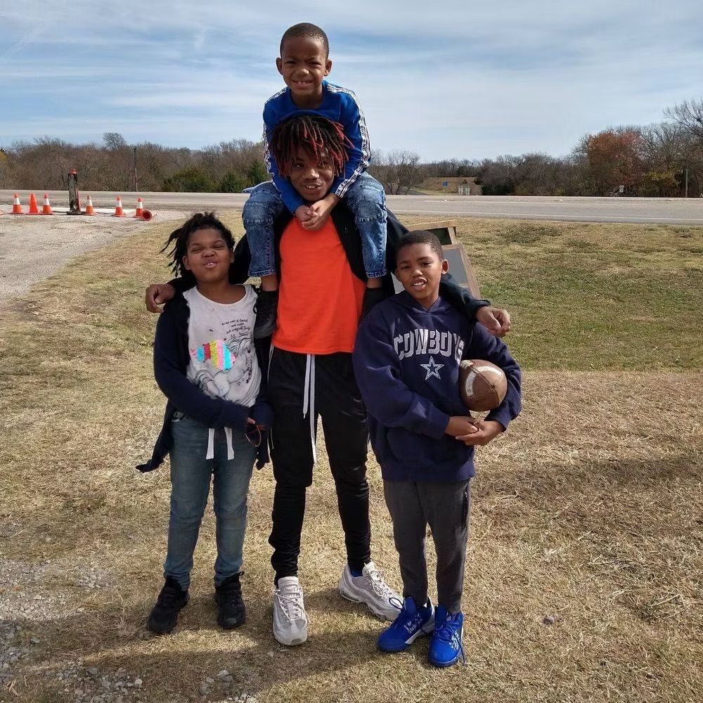 Four children outdoors, one on shoulders, others holding a football, sunny day.