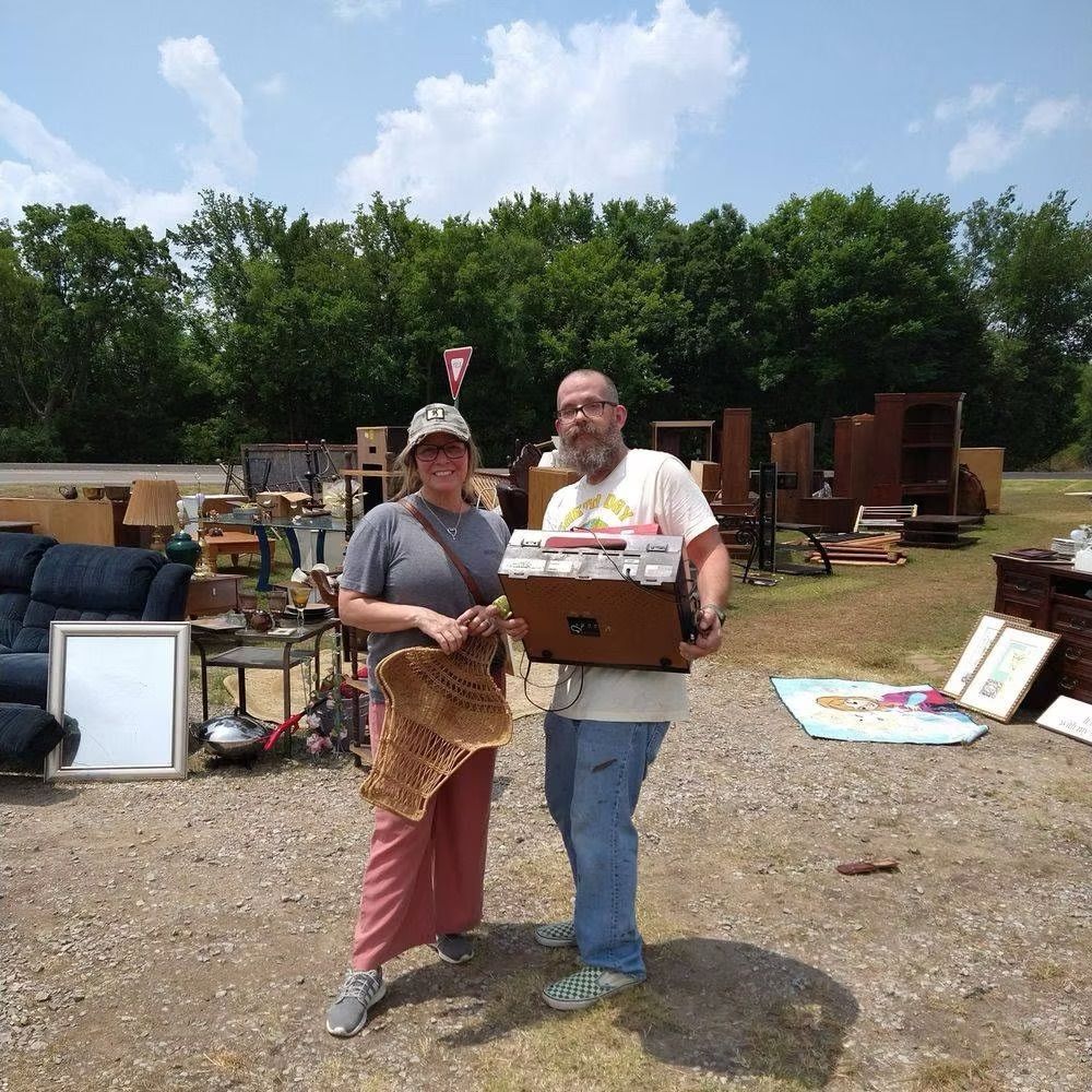 Two people at a yard sale, holding items. Numerous furniture pieces are displayed outside on a gravel lot. Sunny day.