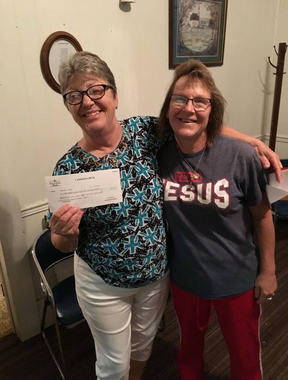 Two women smiling, one holding a check. Interior setting. One has a