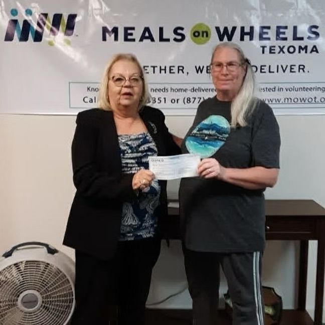 Two women, one in a suit, presenting a check to a woman in a casual shirt, in front of a Meals on Wheels banner.