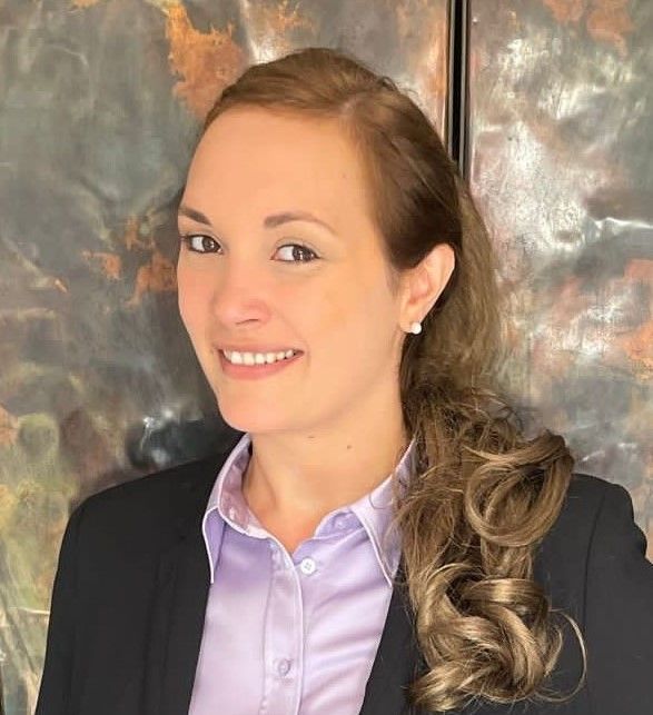 A smiling woman with wavy hair, wearing a lavender collared shirt and black blazer, poses against a textured background.
