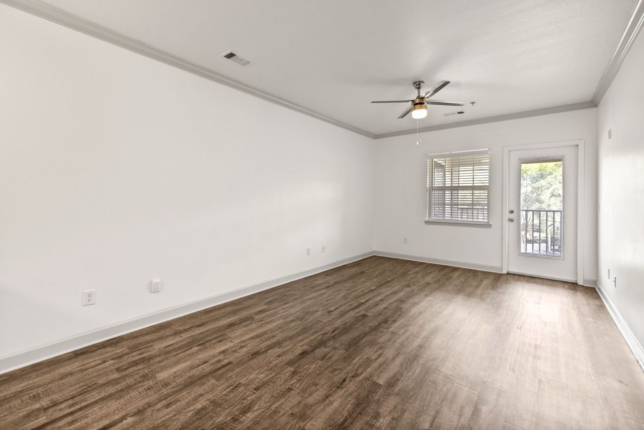 Empty living room with wood-look flooring, white walls, window, ceiling fan, and balcony door.
