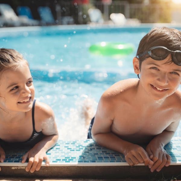 Two children smile while leaning on the edge of a bright blue swimming pool on a sunny day.