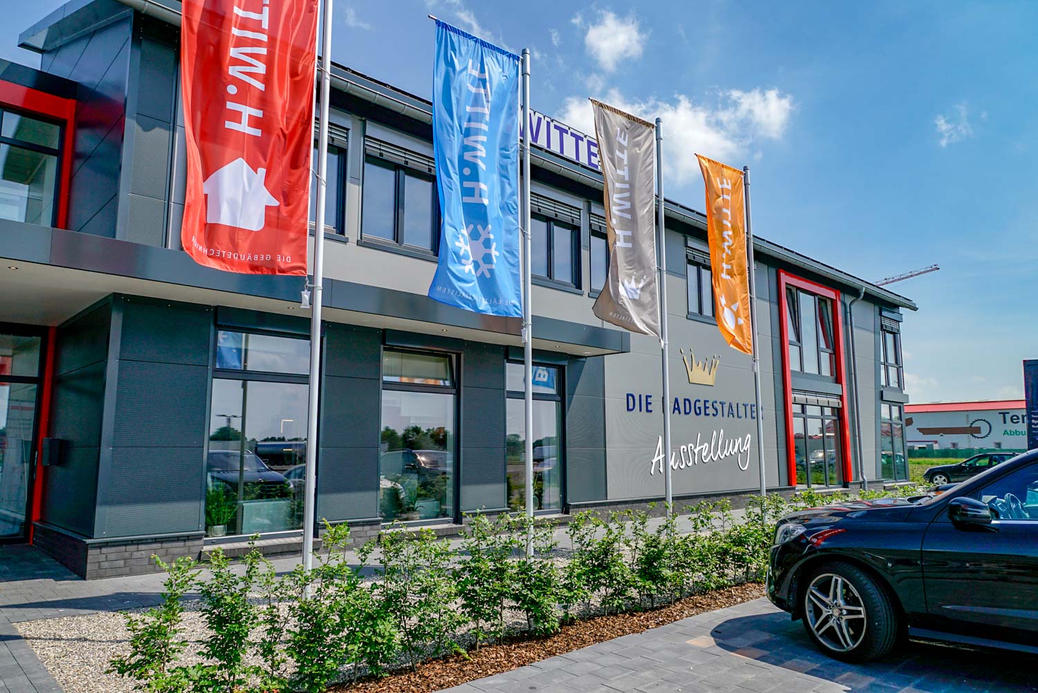 Vor einem Gebäude ist ein Auto geparkt, vor dem Fahnen wehen.