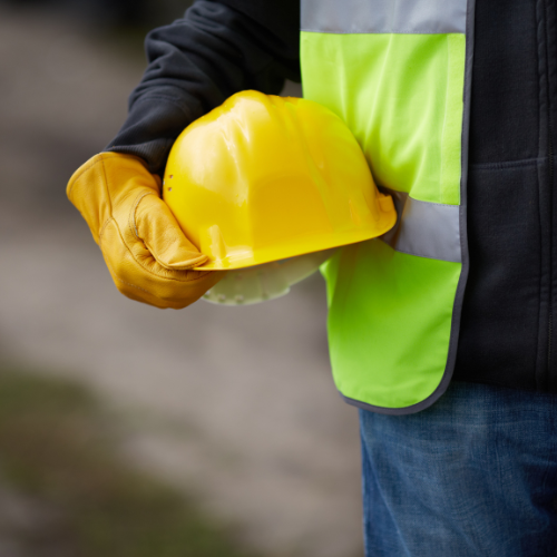 Person in work gear holds a yellow hard hat; a reflective vest and glove are visible.