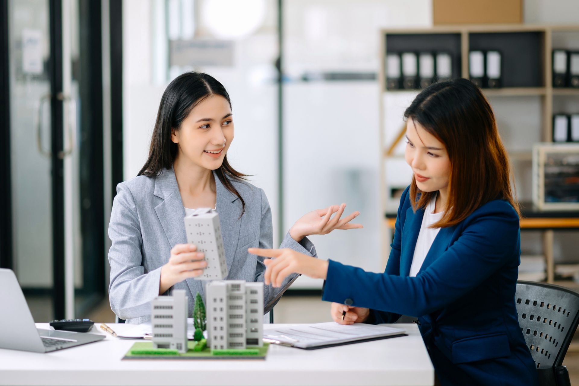 Two women, one pointing at a building model, discussing real estate plans in an office.