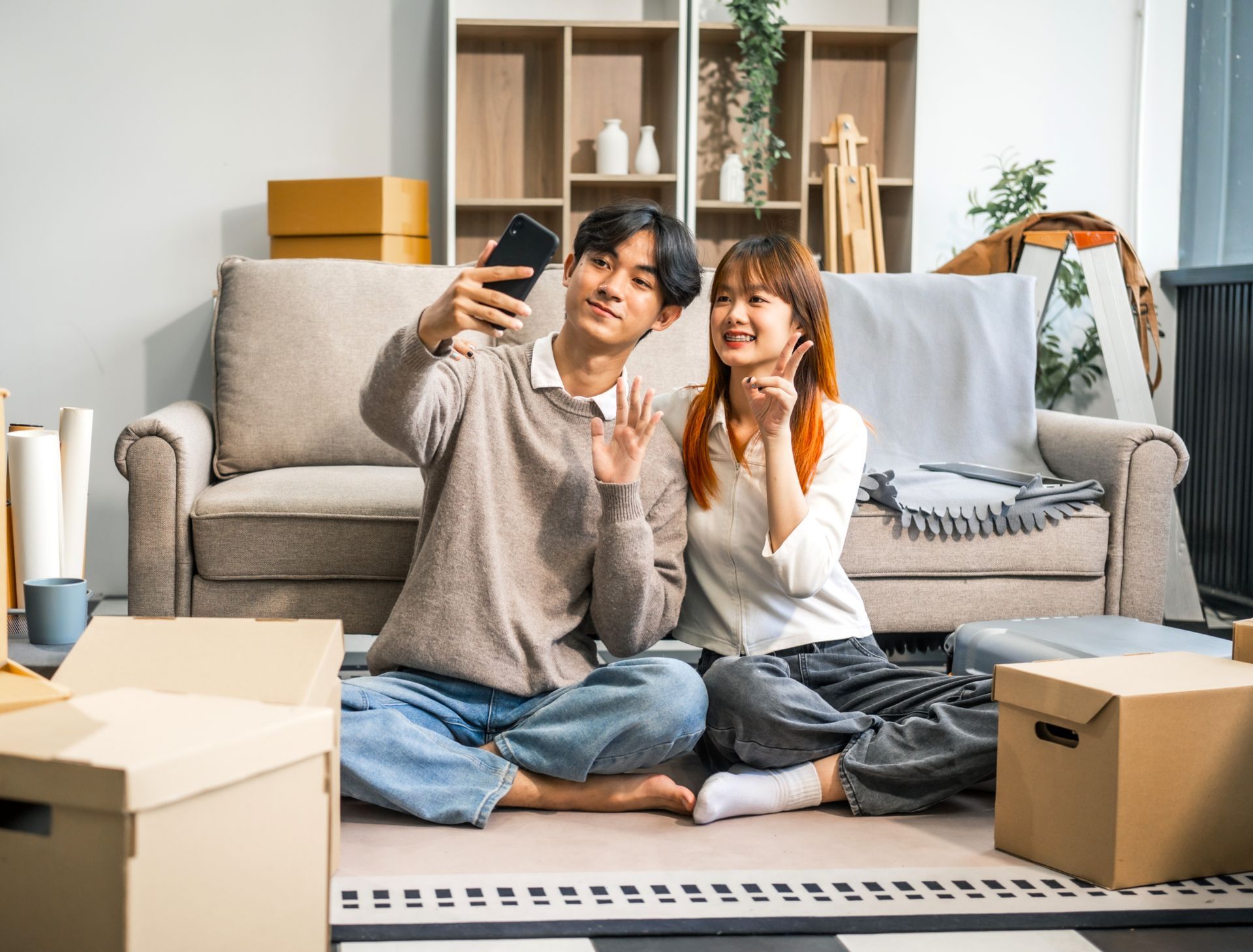 Couple sitting on floor taking a selfie, surrounded by moving boxes in a new home.
