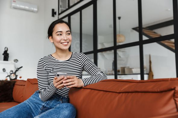 Woman with a smartphone sits on a brown couch, smiling in a modern living room with glass wall.