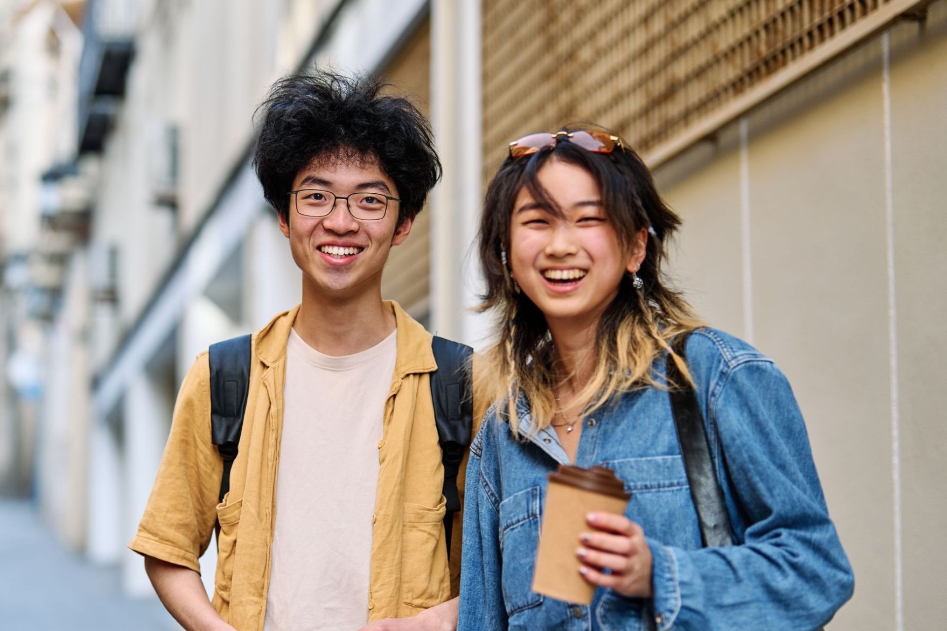 Two young Asian people smile on a city street; the person on the right holds a coffee cup.