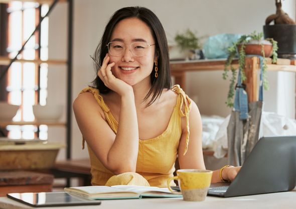 Woman with glasses smiles, resting chin on hand, working on laptop at desk. Bright room, yellow dress.