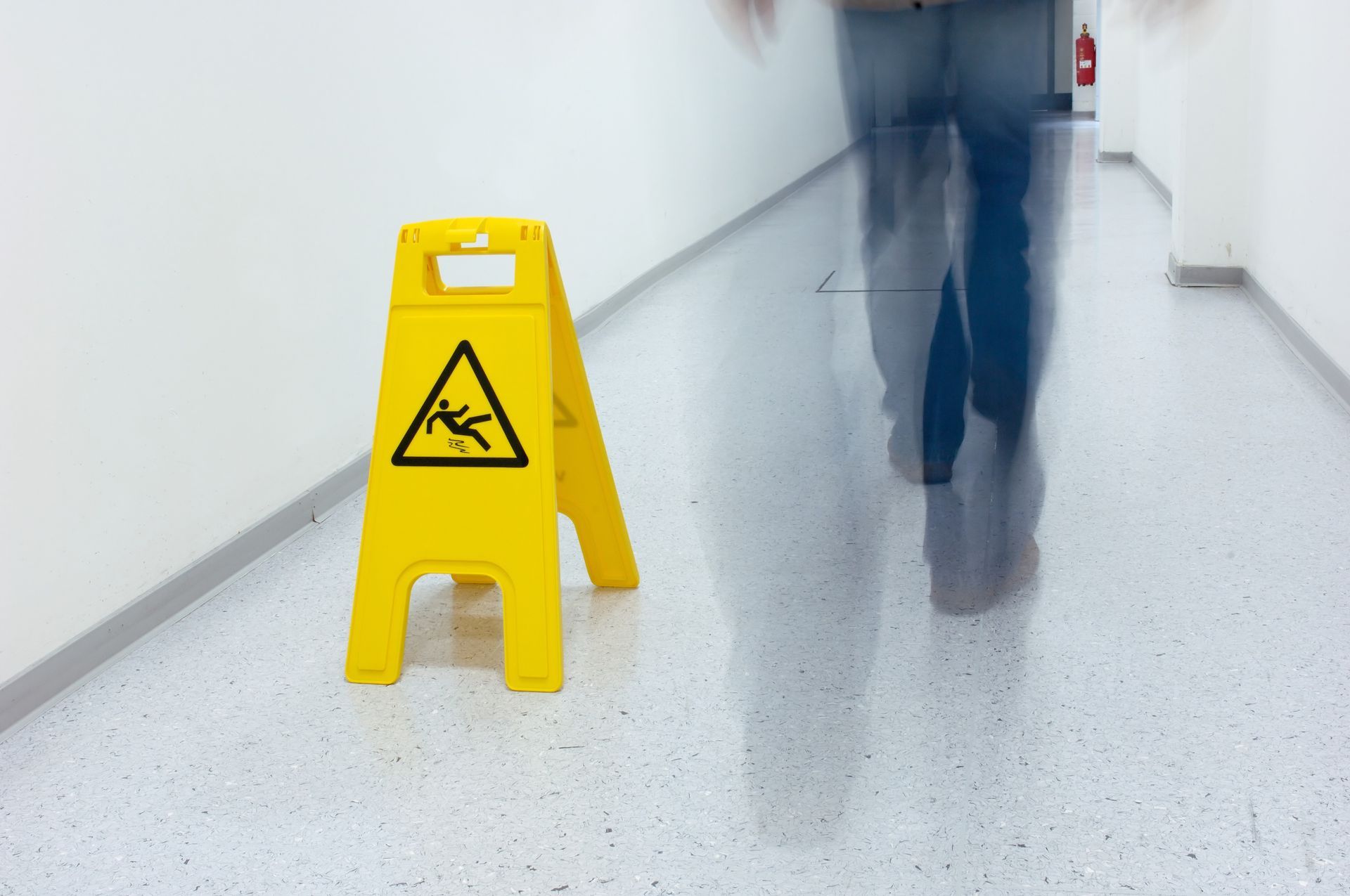 Yellow wet floor sign in a white hallway with a blurred figure walking by.