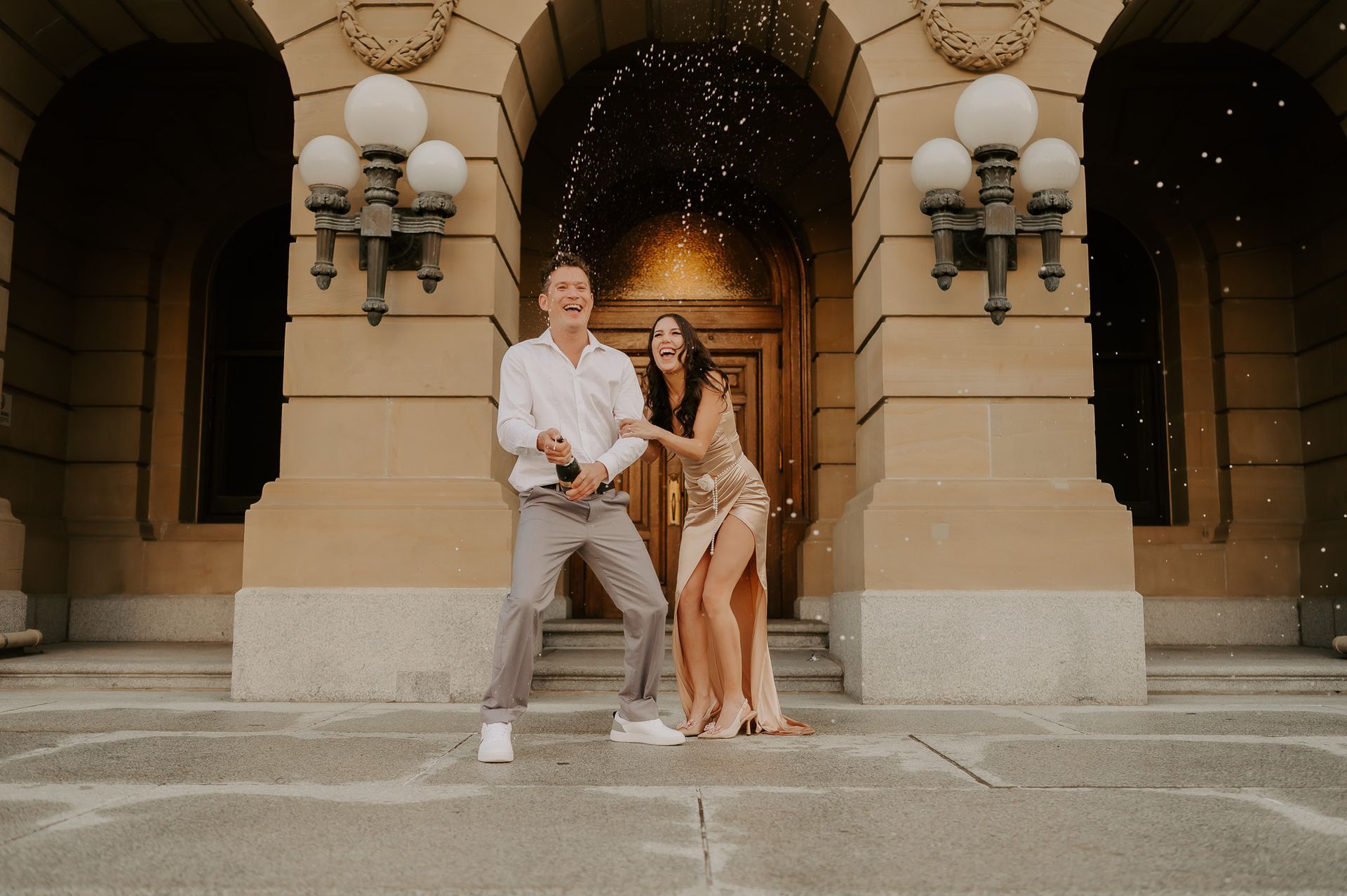 A man and a woman are standing in front of a building holding champagne bottles.