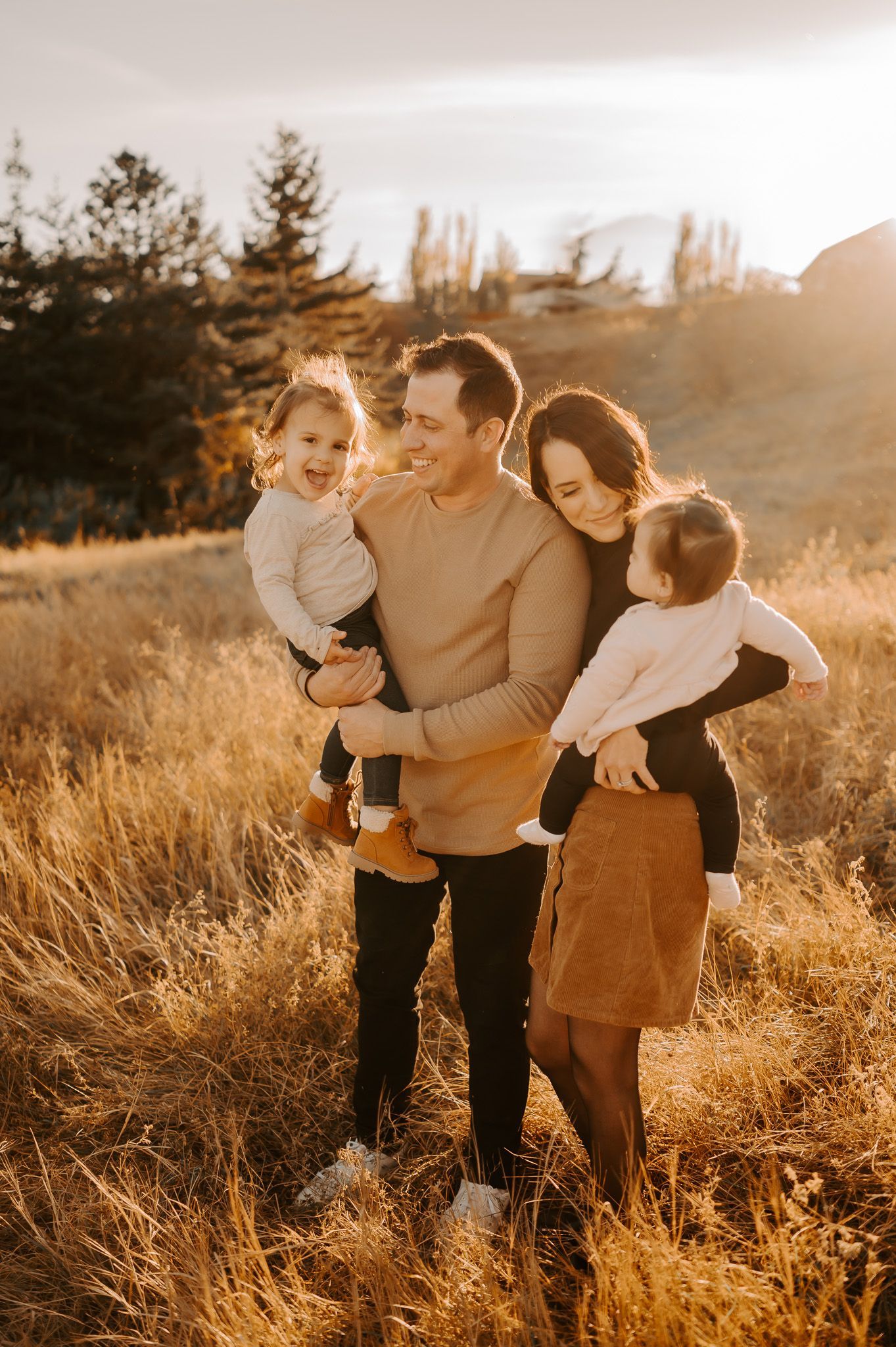 A photo of a family laughing together in a fall setting with a grassy field. Dad is holding sun upside down with sister laughing.