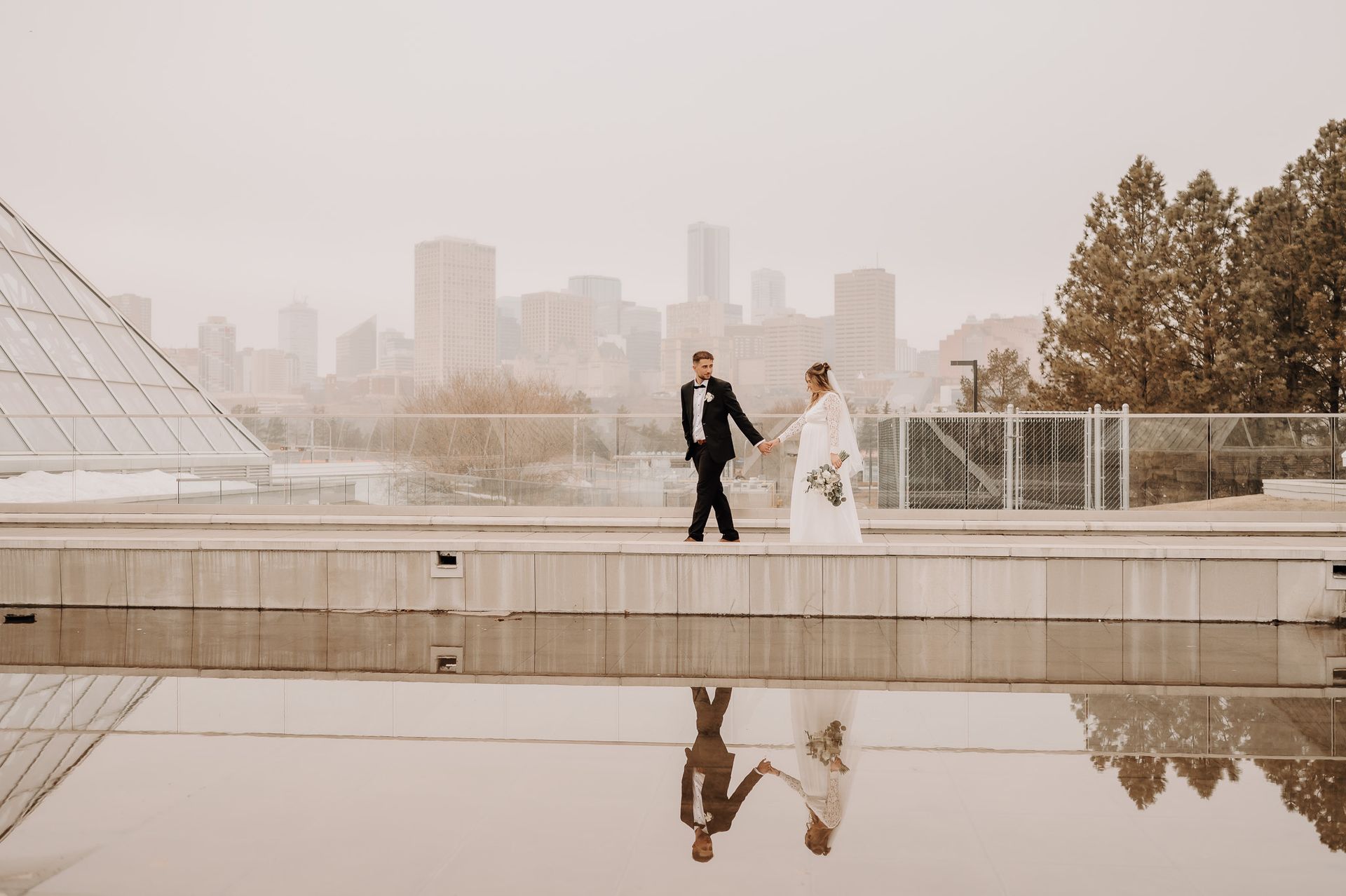 A bride and groom are walking across a bridge in front of a city skyline.