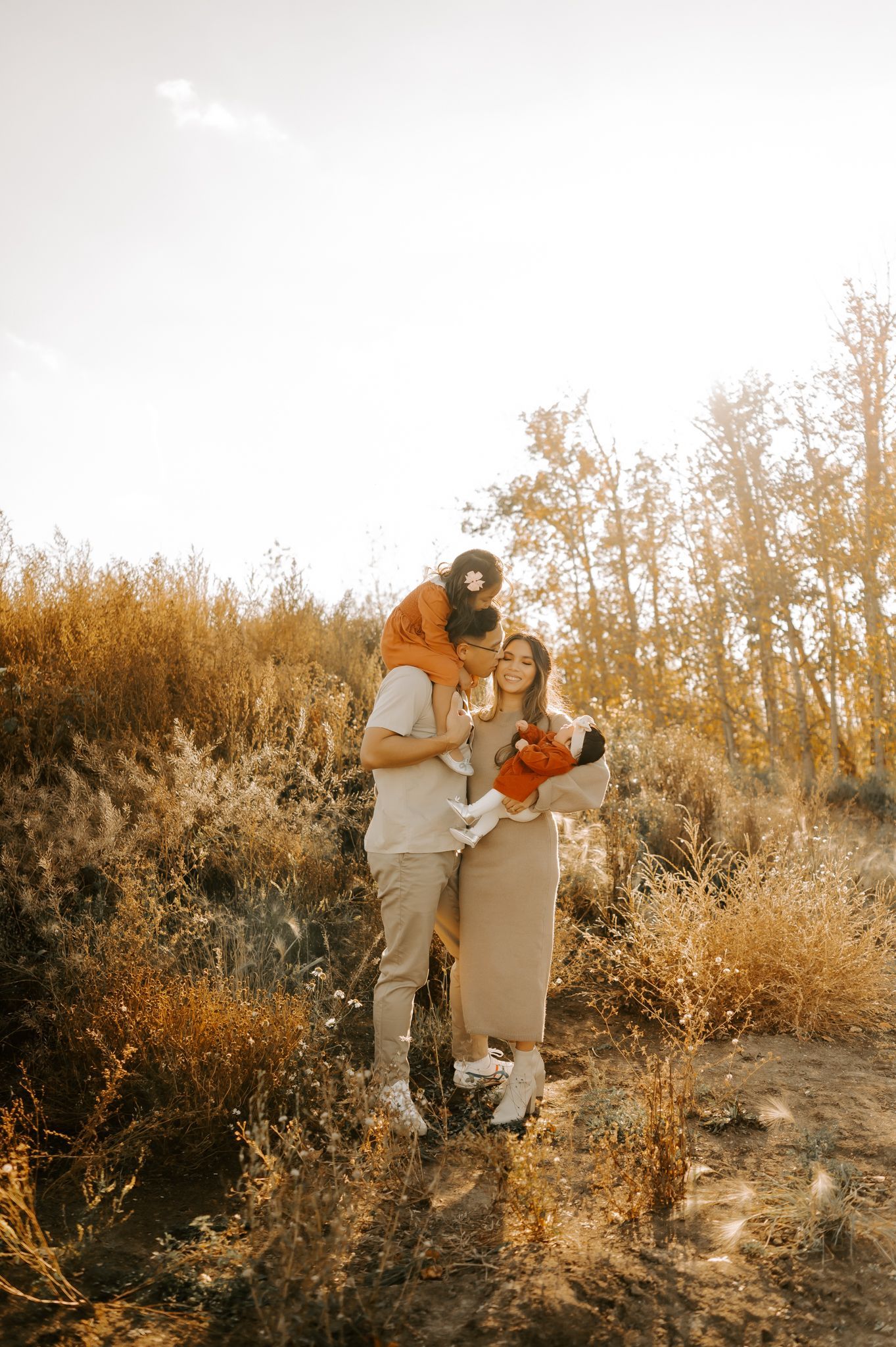 Family photography of a dad kissing the mom on the cheek. His young daughter is on his back, and the mom is holding a baby in a grassy field.