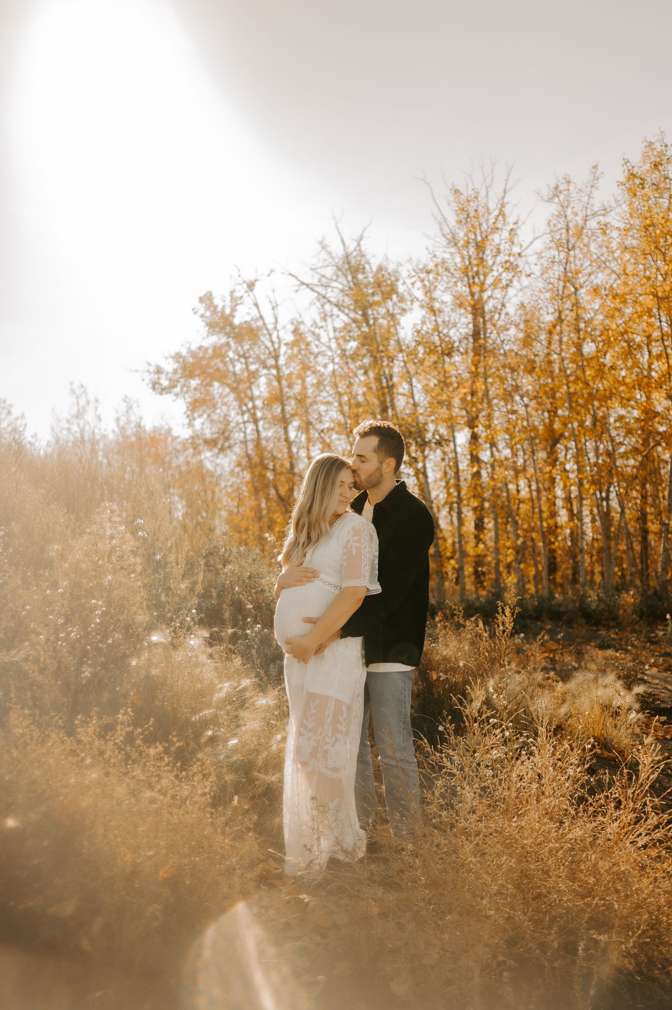 Maternity photography of a mother in a long dress holding her belly. She smiles while looking into the distance, standing in a sandy area.