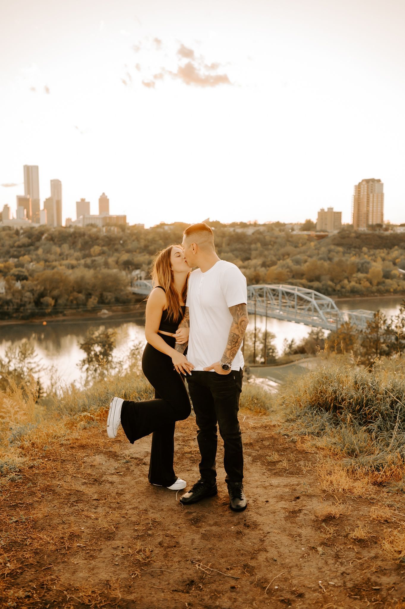 A couple recently engaged holding hands while the woman is on the man's back kissing his cheek as he looks into the distance.