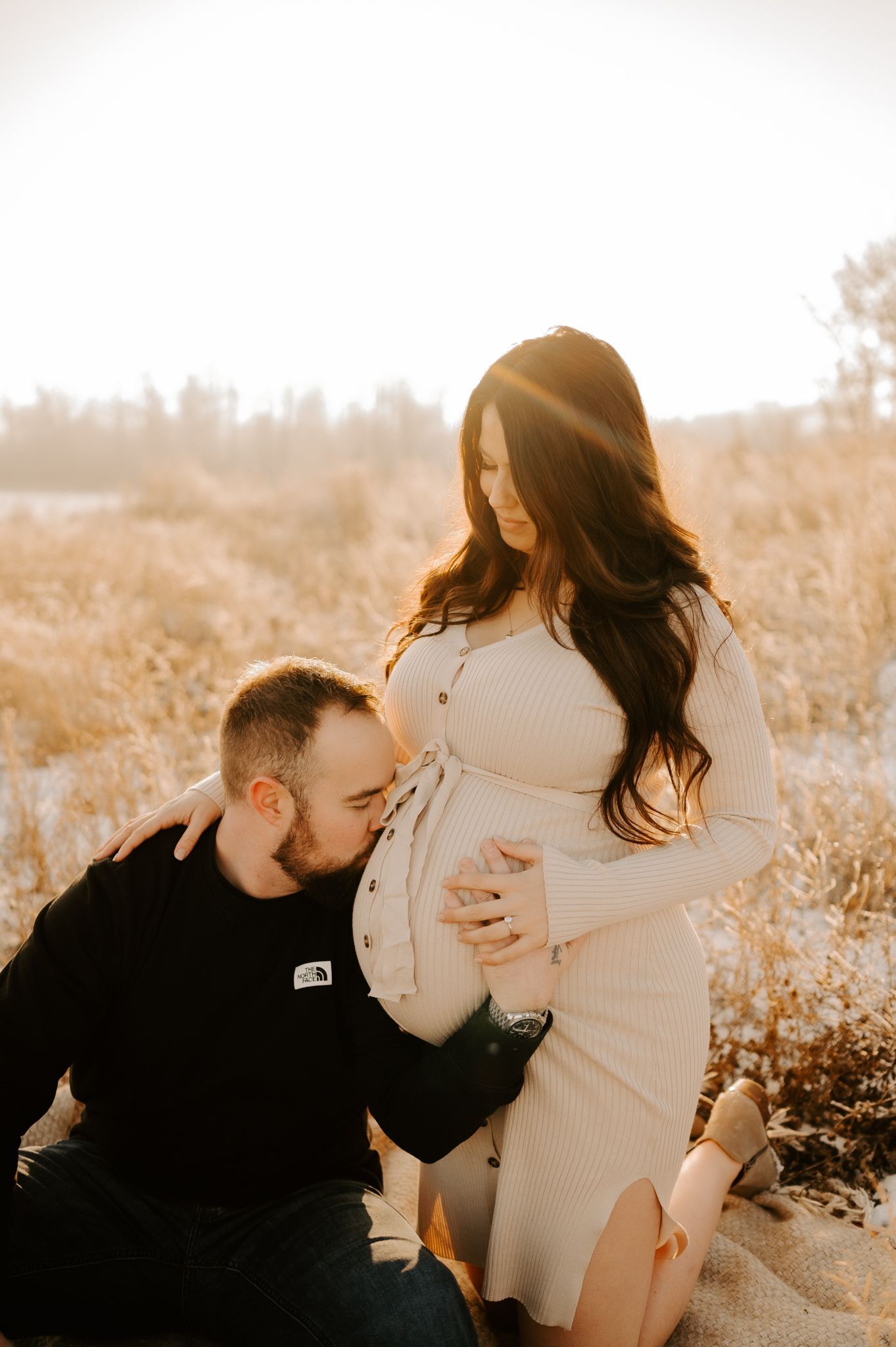 Maternity photography of a mother in a flowing dress in a grassy field, holding her belly and her toddler daughter's hand.