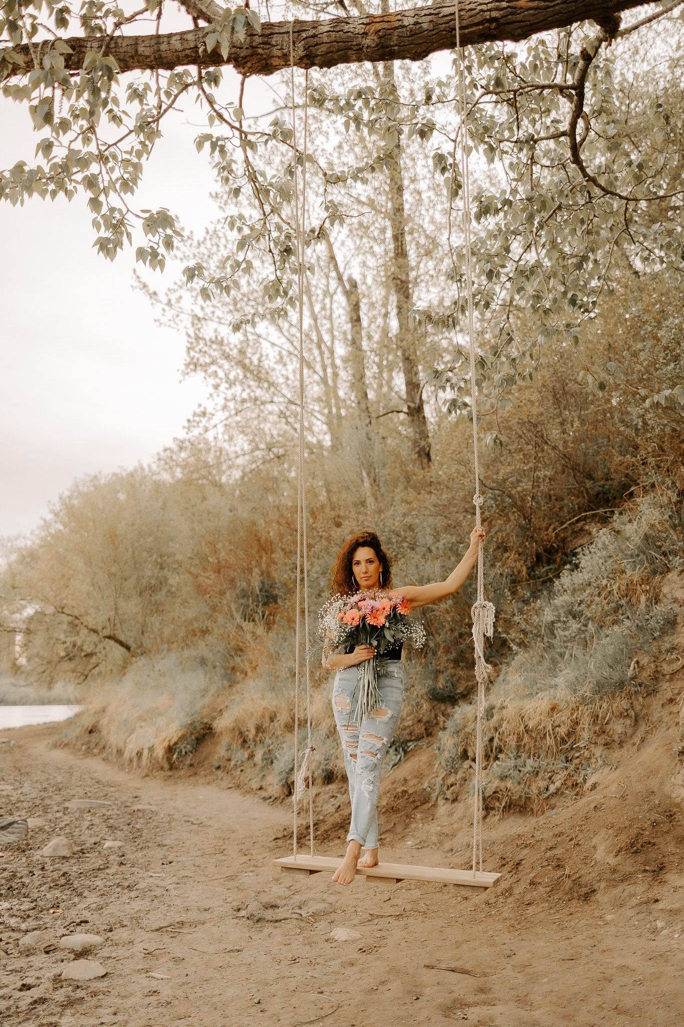 A woman is standing on a swing in the woods holding a bouquet of flowers.