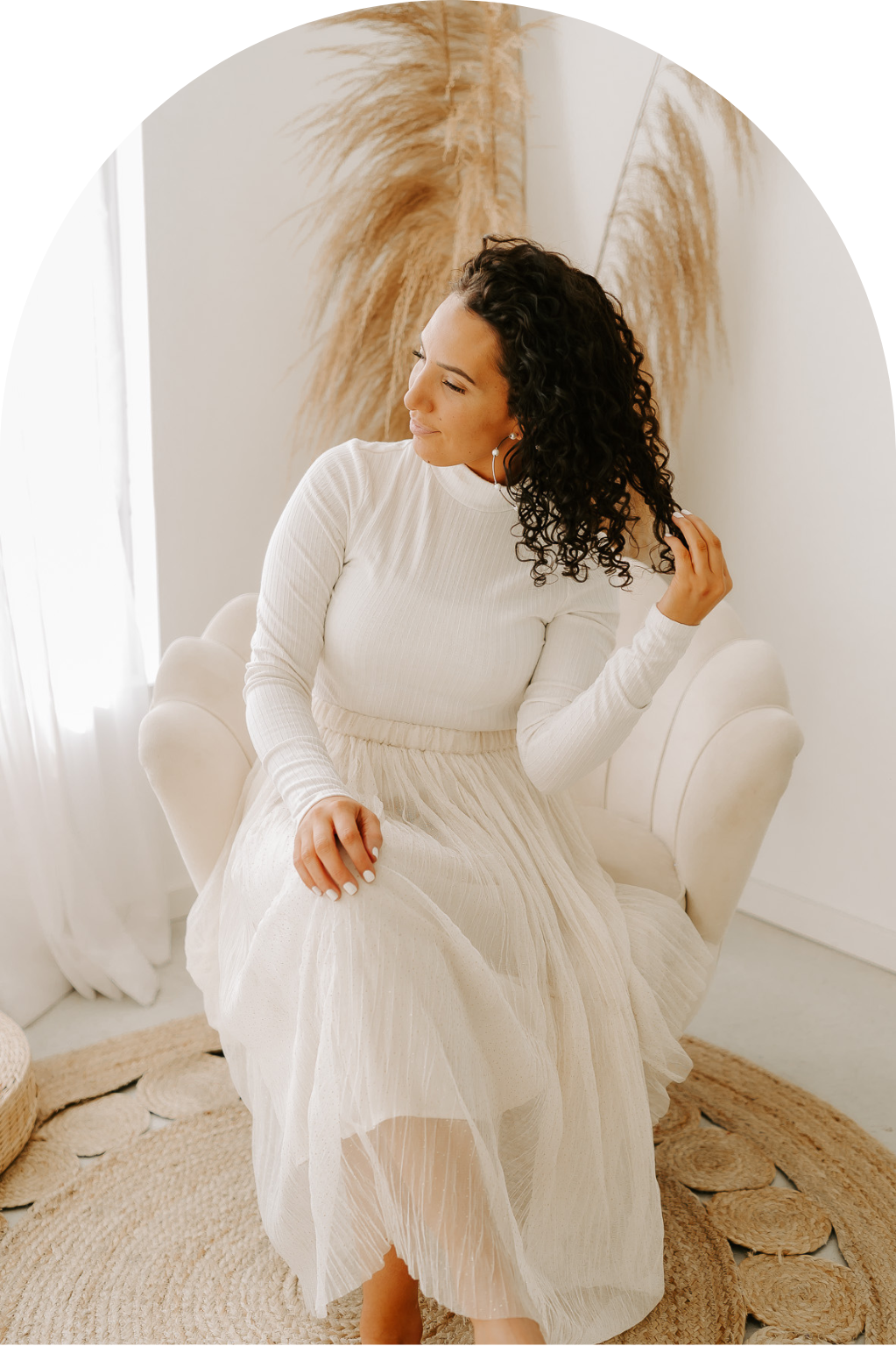 A photo of Edmonton photographer Shayna Fearn Photography, sitting elegantly on a white chair, in a long white dress, playing with her dark curly hair.