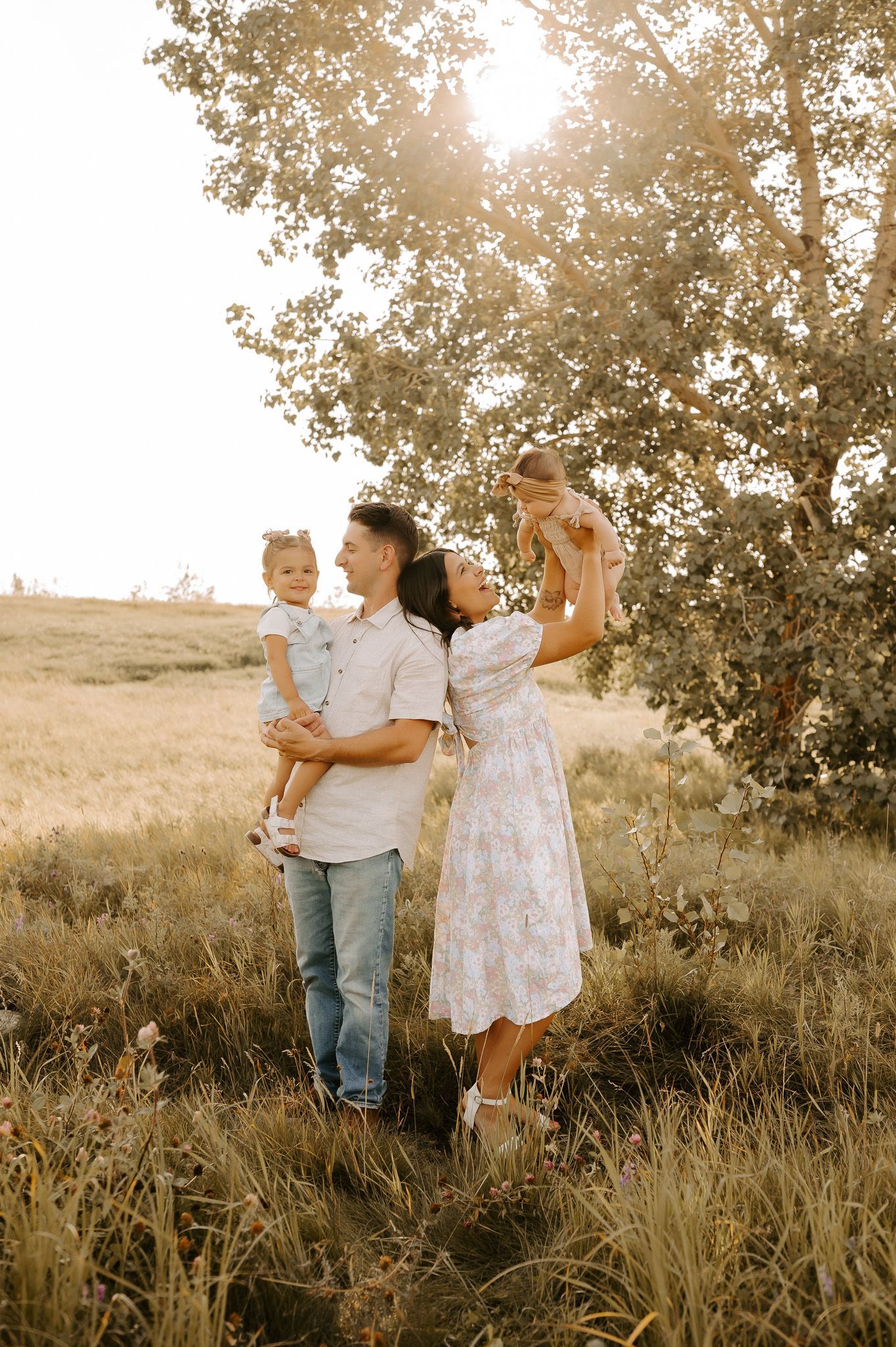 A photo of a family laughing together in a fall setting with a grassy field. Dad is holding sun upside down with sister laughing.