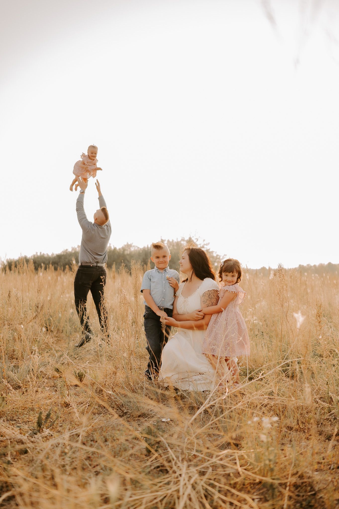 A photo of mom and dad kissing in a golden sunset with autumn trees in the back, the baby son is on the dads shoulders.