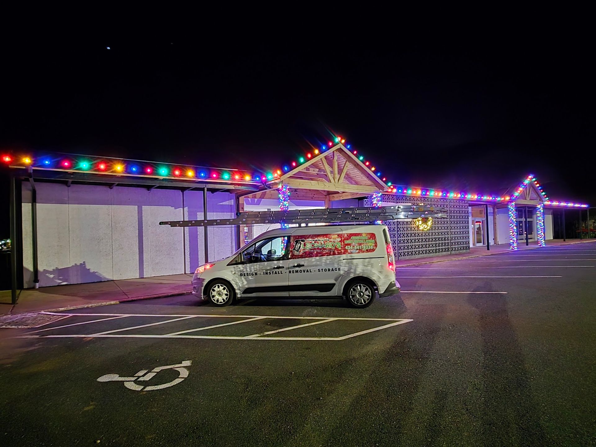 A silver van parked in front of a building decorated with colorful Christmas lights at night.