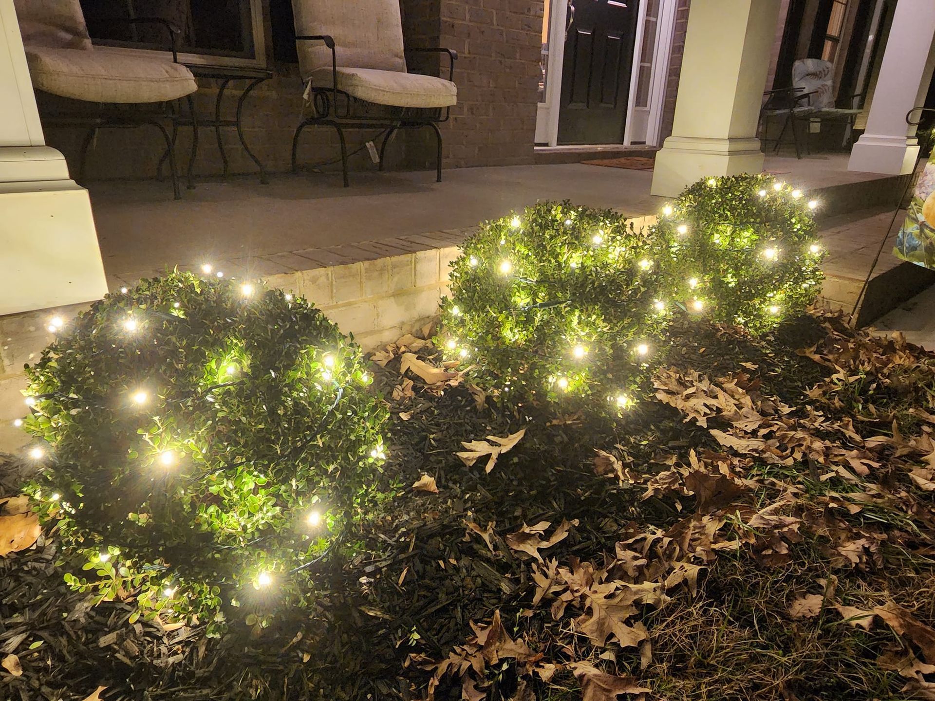 Three round bushes decorated with glowing white lights, in front of a porch.