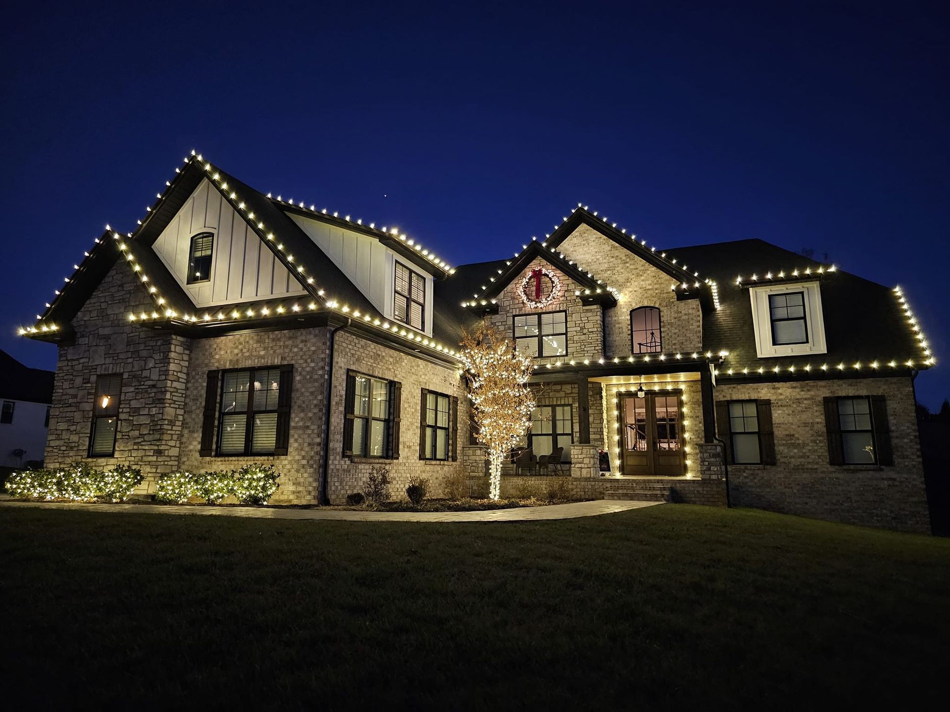 Two-story brick house illuminated with white Christmas lights outlining the roof, windows, and front door at night.