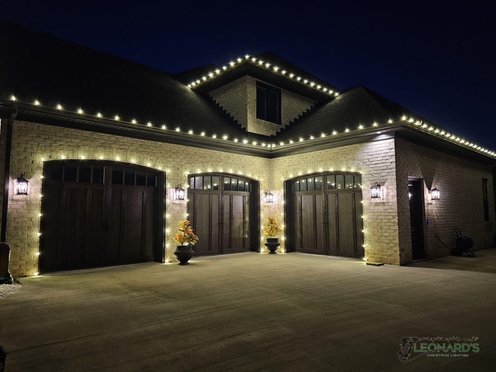 House exterior at night, decorated with white Christmas lights along rooflines and garage doors.