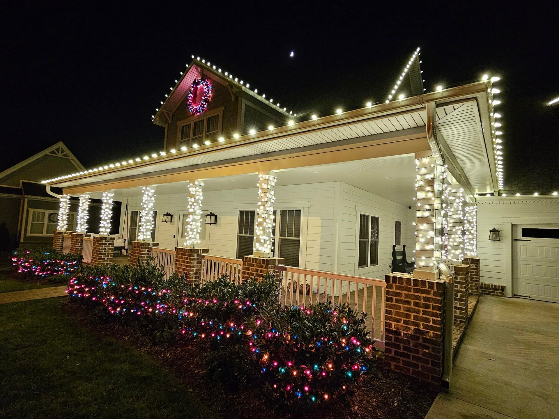 House decorated with Christmas lights at night; white house with lights outlining roof, columns, bushes, and a wreath.