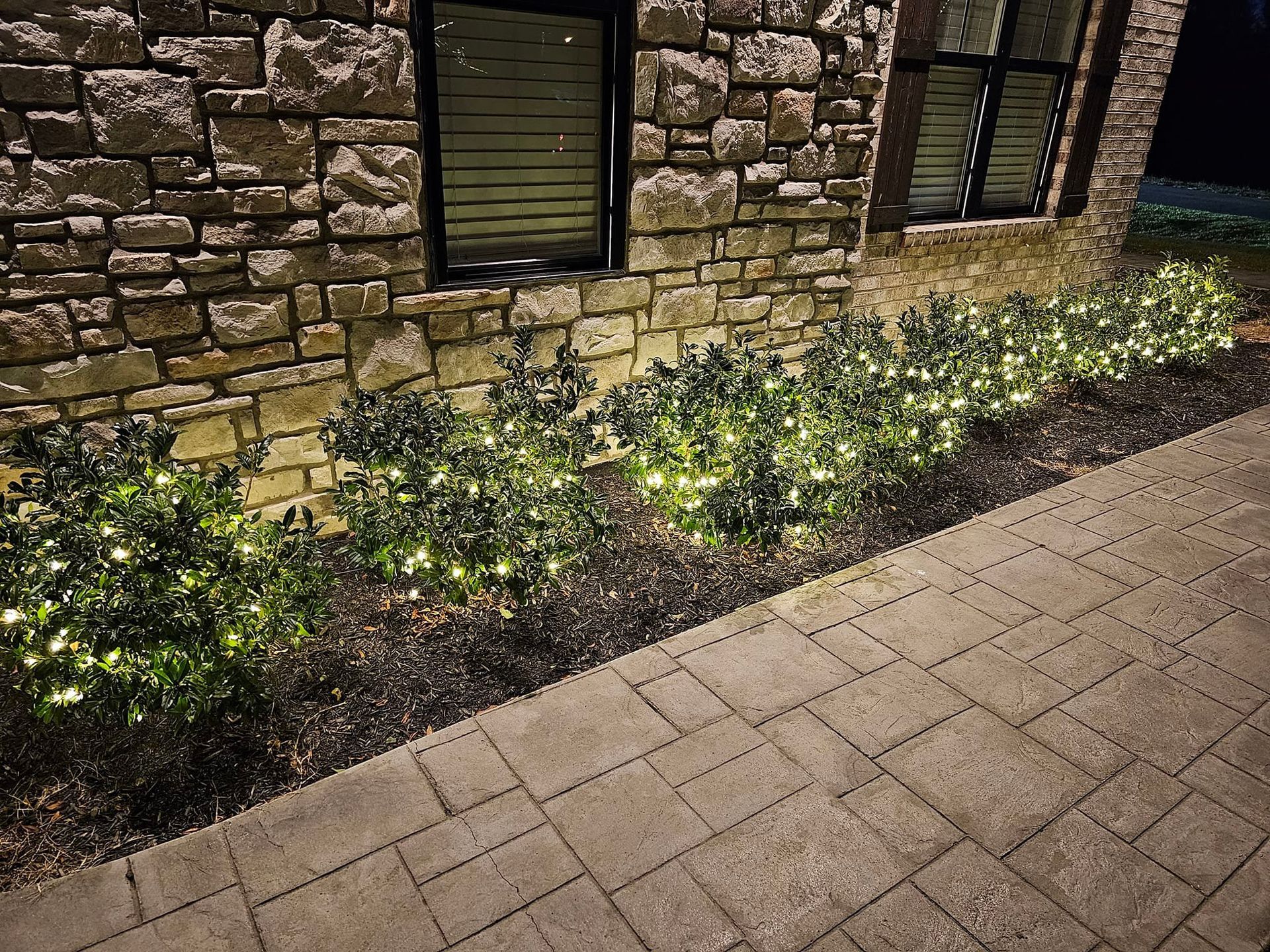 Stone building with illuminated bushes in a dark setting, next to a paved pathway.