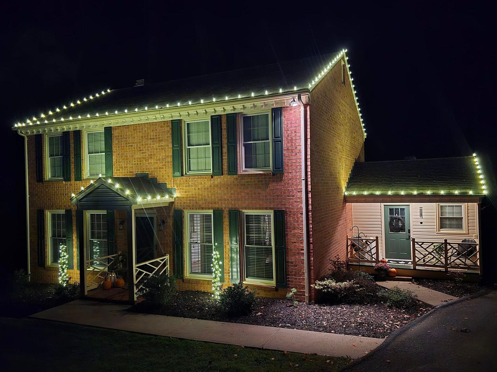 Brick house with green shutters and Christmas lights, a small building is next to it with a porch.