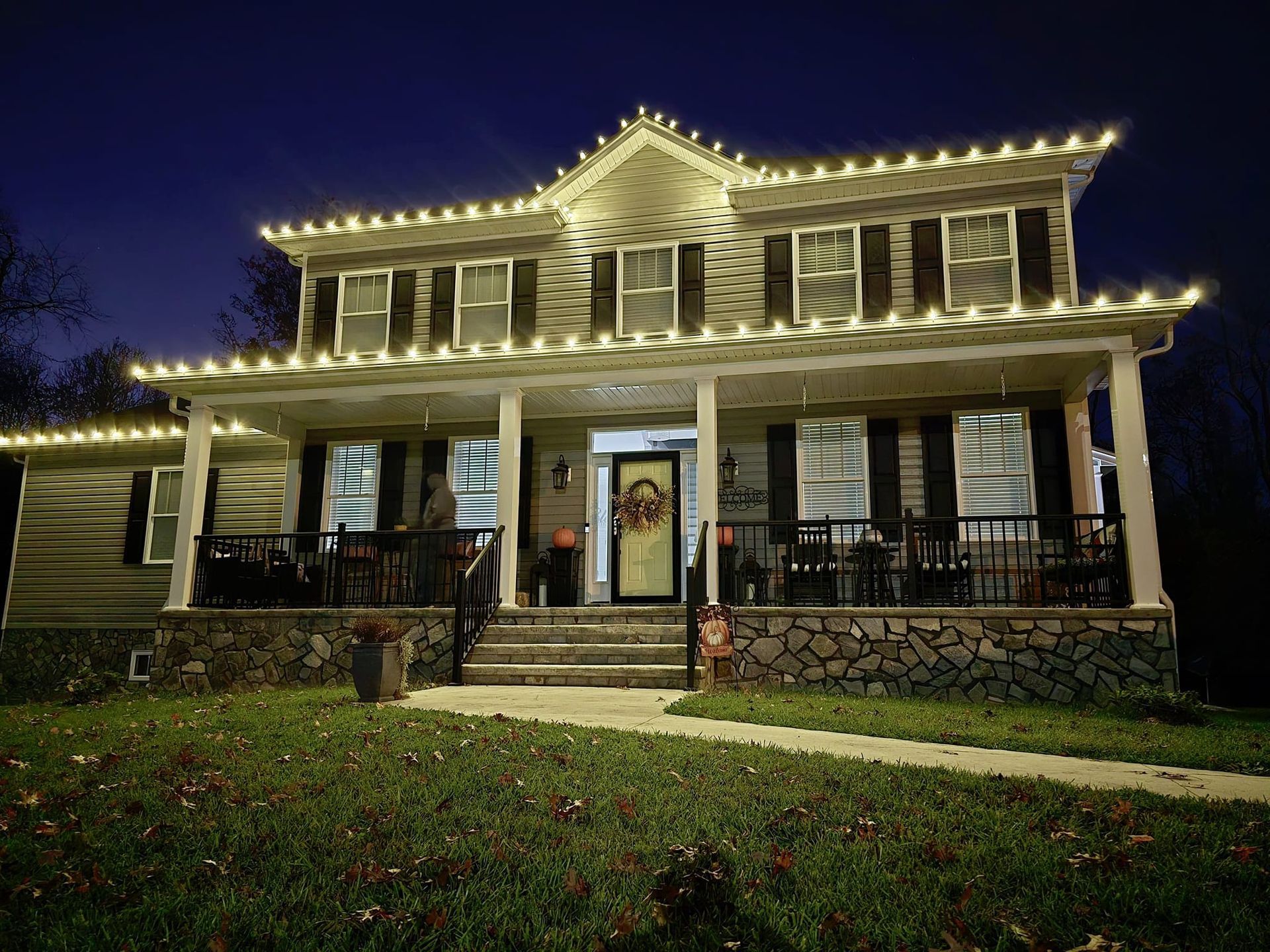 Two-story house at night, decorated with white lights along the rooflines and porch.