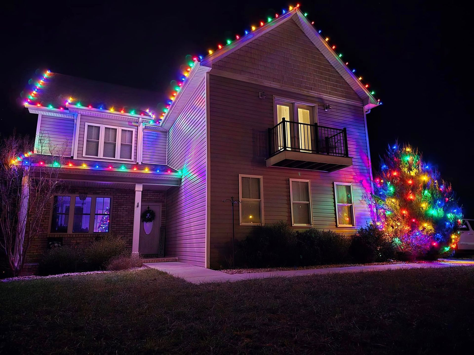 Two-story house lit with colorful Christmas lights at night, with a lit tree in front.
