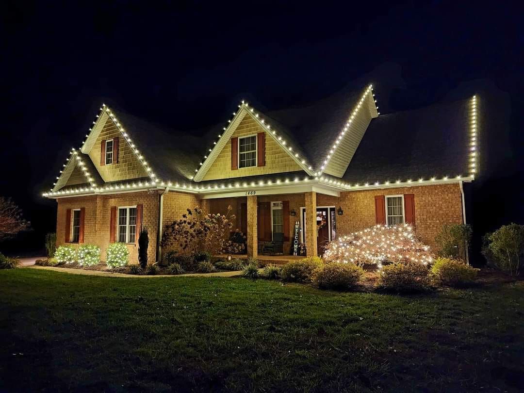 House at night with roof outlined in white lights, lit bushes, and a green lawn.