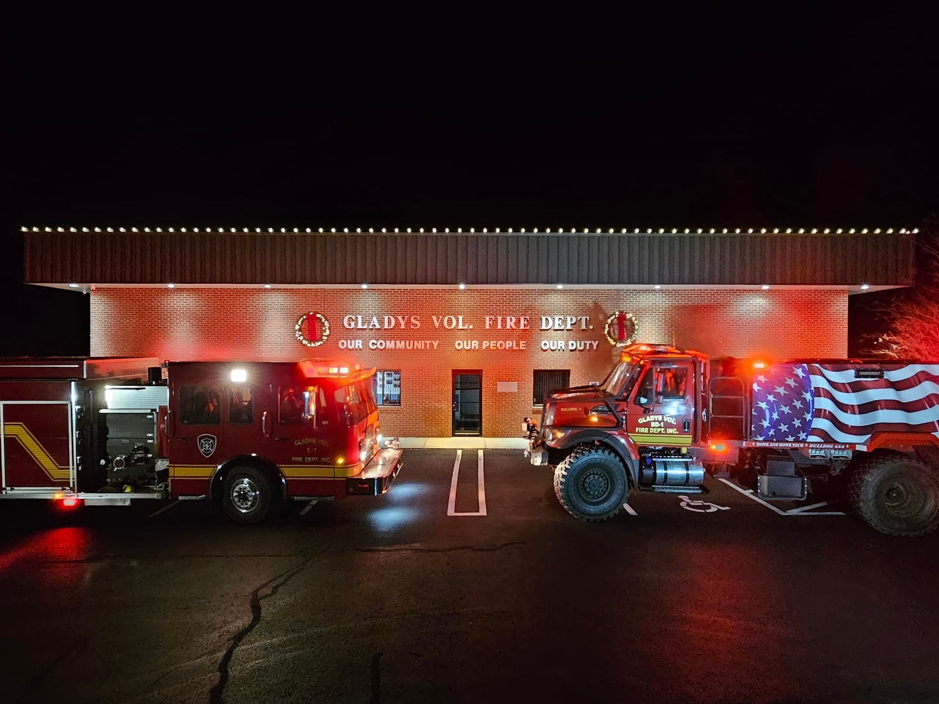 Fire trucks parked in front of fire station at night, lights on. One truck bears a draped flag.