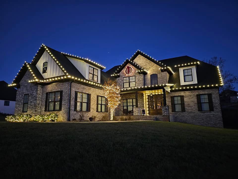 House at dusk with white Christmas lights on the roof and a tree in front, on a grassy lawn.